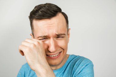 Ağlayan genç adamın yüzü, elleri ve ağzı açık açık, açık arkaplanda mavi bir tişört giymiş..