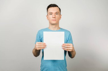 Mavi tişörtlü genç adam arka planda bir sayfa A4 tutuyor..