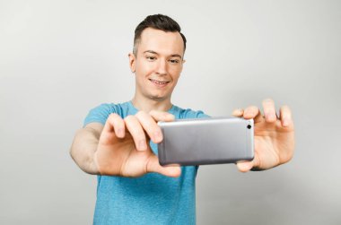 Mavi tişörtlü komik beyaz adam açık gri arka planda akıllı telefon kamerasıyla özçekim yapıyor..