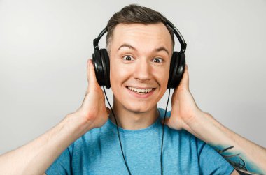 Mavi tişörtlü komik beyaz adam açık gri arka planda kulaklıkla müzik dinliyor..