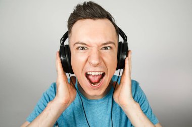 Mavi tişörtlü komik beyaz adam açık gri arka planda kulaklıkla müzik dinliyor..