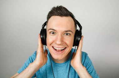 Mavi tişörtlü komik beyaz adam açık gri arka planda kulaklıkla müzik dinliyor..