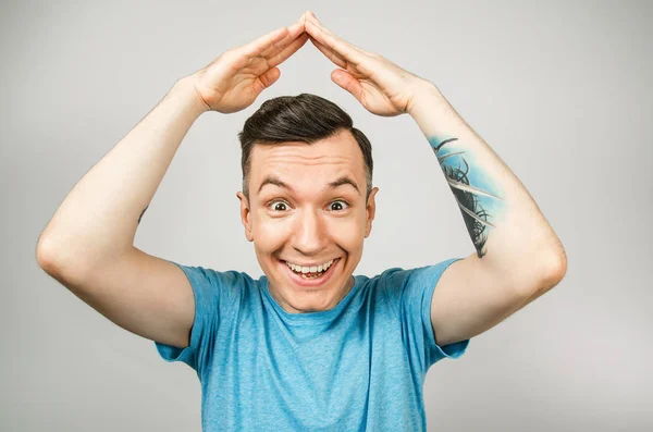 Jeune homme souriant vêtu d'un t-shirt bleu sur fond clair. Le gars tient ses mains sur sa tête en forme de toit de la maison — Photo