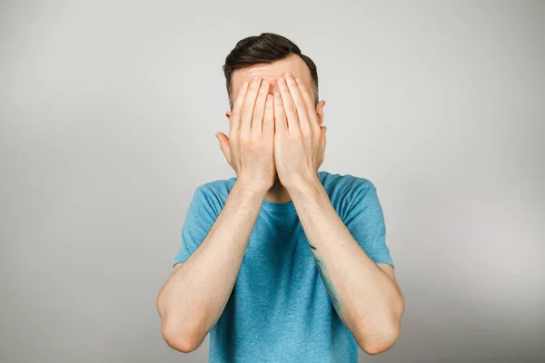 Young smile guy dressed in a blue t shirt, close his eyes with hands on a light background. — 스톡 사진