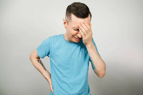 Νεαρός τύπος που κλαίει καλύπτει το πρόσωπο με τα χέρια με ανοιχτό το στόμα, ντυμένος με ένα μπλε t πουκάμισο σε ένα ελαφρύ φόντο. — Φωτογραφία Αρχείου