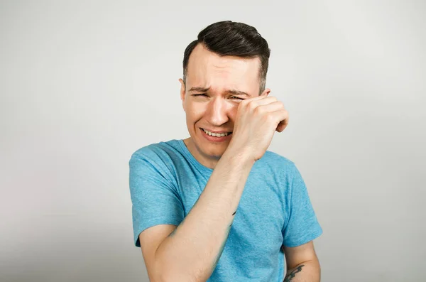 Young crying guy cover face with hands with open mouth, dressed in a blue t shirt on a light background. — Stock Photo, Image