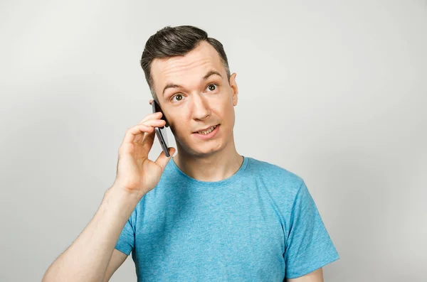 Νεαρός θυμωμένος καυκάσιος με μπλε μπλουζάκι μιλάει στο τηλέφωνο, σε ανοιχτό γκρι φόντο. — Φωτογραφία Αρχείου