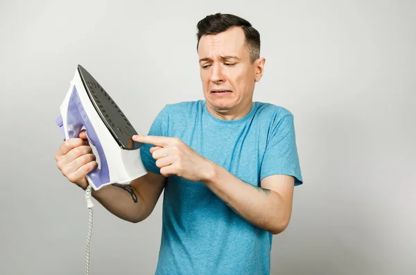 The young guy get burnt with fear keeps a smoothing iron and checks the temperature with his finger on a light gray background. — Stock Photo, Image