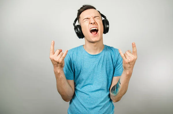 Młody, zabawny biały facet w niebieskiej koszulce słuchający muzyki na słuchawkach na jasnoszarym tle. — Zdjęcie stockowe