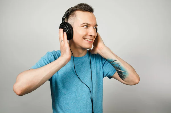 Ung rolig kaukasier kille i blå t-shirt lyssna på musik på hörlurar på ljusgrå bakgrund. — Stockfoto