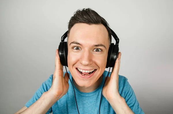 Joven chico caucásico divertido en camiseta azul escuchando música en los auriculares sobre fondo gris claro . —  Fotos de Stock