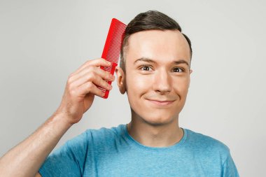 Genç adam bir tarak tutuyor ve saçını tarıyor ve beyaz bir arka planda gülümsüyor. Yakın plan portre.