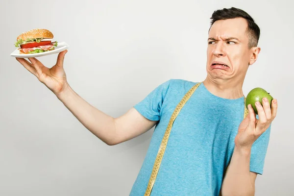 Mladý usmívající se muž s měřicí páskou zavěšenou na ramenou, drží hamburger a zelené jablko, na bílém pozadí. Koncept výběru mezi zdravými a nezdravými potravinami — Stock fotografie