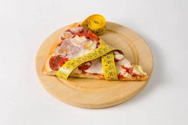 Piece of pizza wrapped in a measuring tape lies on a round wooden board on a white background. Top view. — 스톡 사진