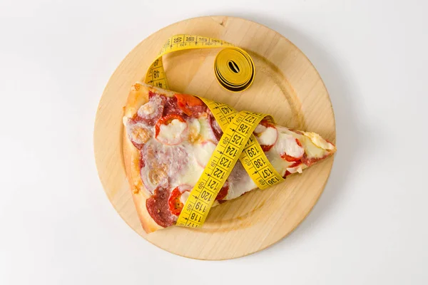 Pedaço de pizza envolto em uma fita métrica encontra-se em uma tábua de madeira redonda em um fundo branco. Vista superior . — Fotografia de Stock