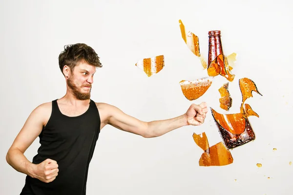A young bearded guy on a white background beats his fist on a glass bottle of beer and it flies to pieces. Concept of fighting alcohol. — Stock Photo, Image