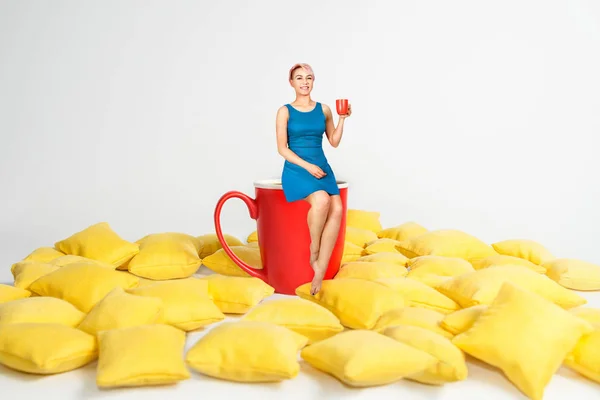 Fiatal gyönyörű lány kék ruhában ül egy hatalmas csésze sárga párnák és tartja csésze kávé fehér alapon. — Stock Fotó