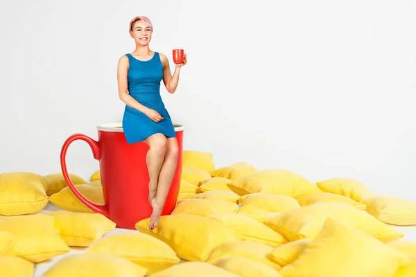 Young beautiful girl in blue dress sits on a huge cup in a yellow pillows and holds cup of coffee on a white background. — Stock Photo, Image