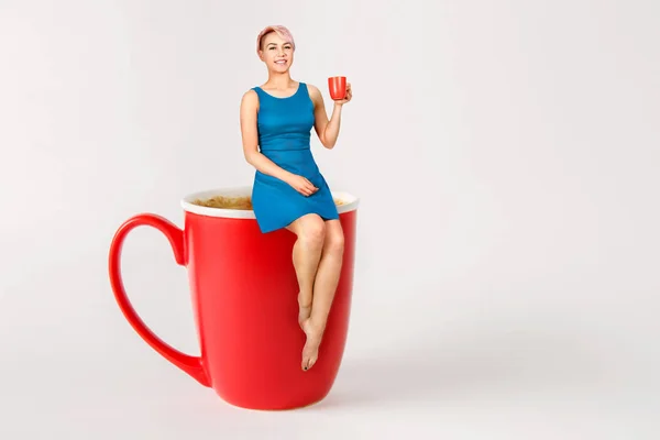 Junges schönes Mädchen sitzt auf einer großen Tasse Kaffee und trinkt Kaffee auf hellem Hintergrund. — Stockfoto