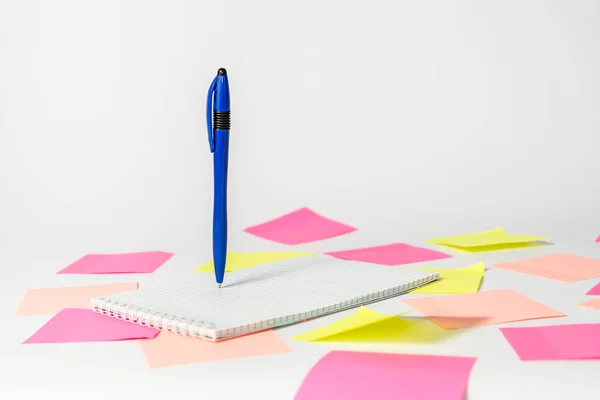 Růžové a žluté samolepky pod zápisníkem, na kterém je napsáno pero, které visí ve vzduchu na světlém pozadí. — Stock fotografie
