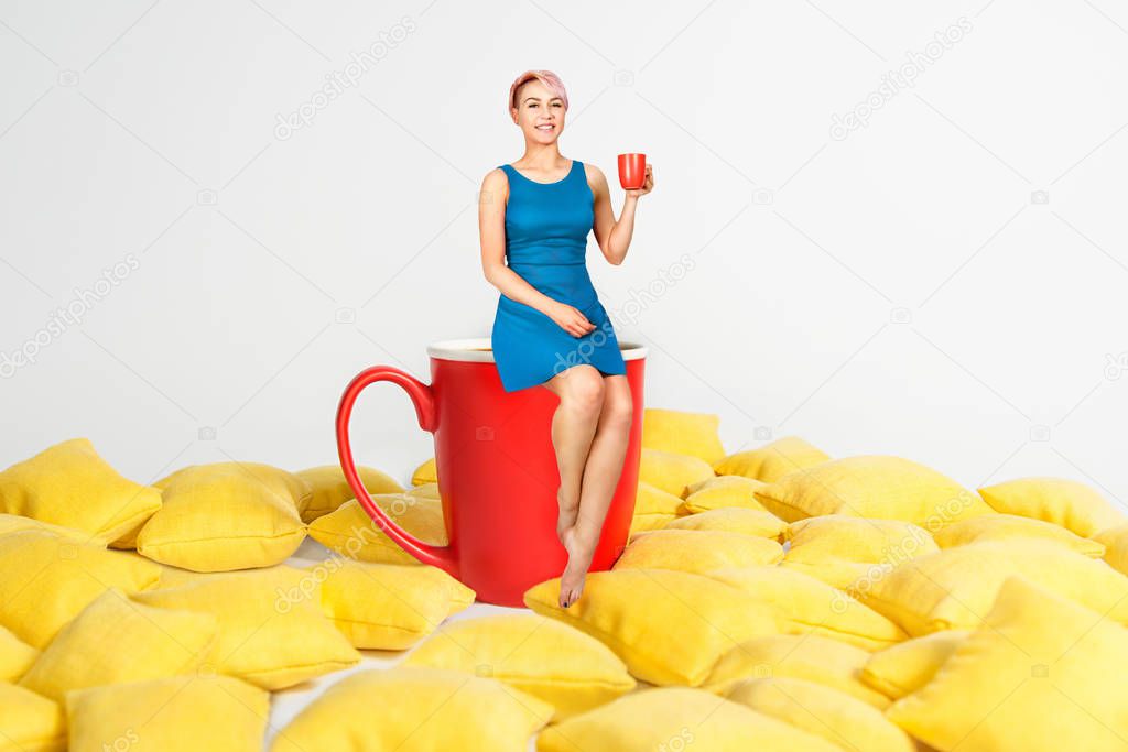 Young beautiful girl in blue dress sits on a huge cup in a yellow pillows and holds cup of coffee on a white background.