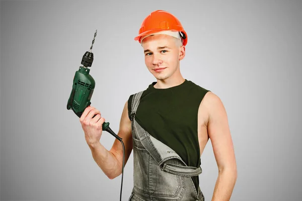 Young working guy in hard hat holds drill on a gray background — Stock Photo, Image