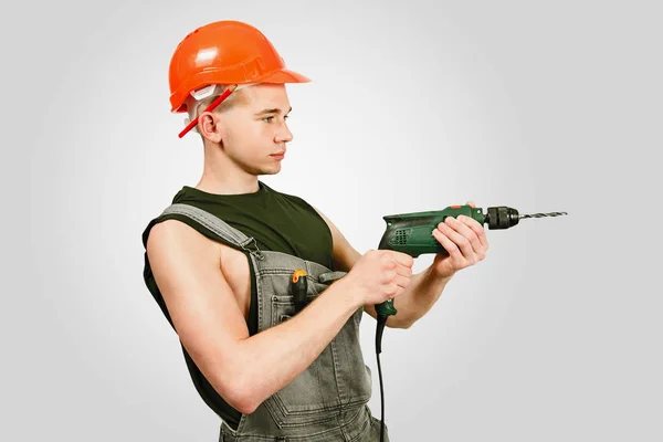 Young working guy in hard hat holds drill on a gray background — Stock Photo, Image