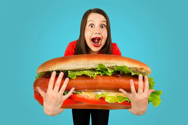 Young cute funny girl holds in front of herself huge hot dog on a blue background — 스톡 사진