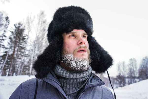 Siberian Russian man with a beard in hoarfrost in freezing cold in the winter freezes and wears a hat with a earflap.