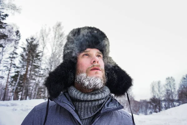 Hombre ruso siberiano con barba en la escarcha en el frío helado en el invierno se congela y lleva un sombrero con una orejera . — Foto de Stock