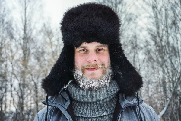 Hombre ruso siberiano con barba en la escarcha en el frío helado en el invierno se congela y lleva un sombrero con una orejera . — Foto de Stock