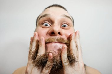 Stüdyoda gri bir arka planda tek başına dikilirken sakalını tarak ile tarayan genç bir adamın yakın çekimi.