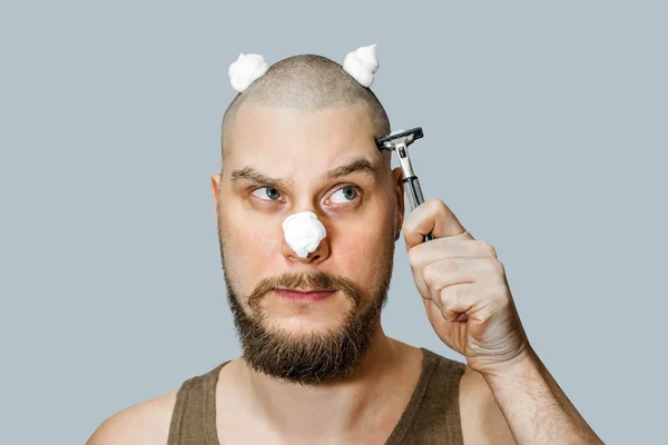Un tipo con barba y una navaja en la mano, con espuma y gel en la cara se afeita por la mañana en casa —  Fotos de Stock