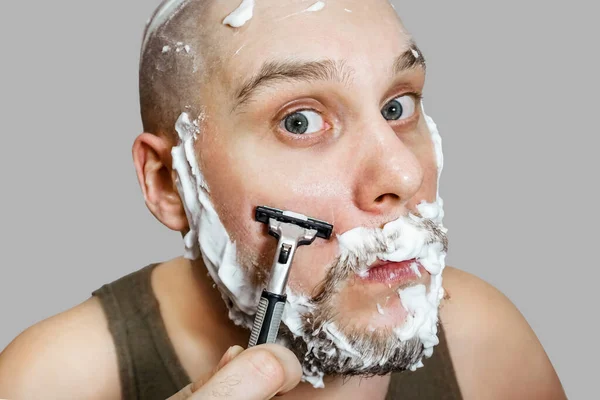 En kille med skägg och rakhyvel i handen, med skum och gel på ansiktet rakar sig på morgonen hemma — Stockfoto