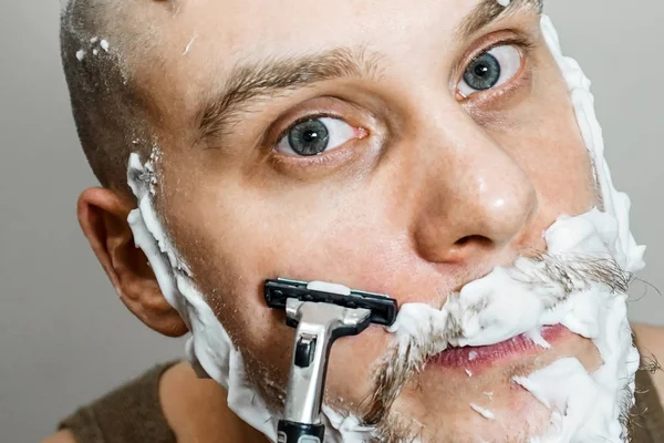 A guy with a beard and a razor in his hand, with foam and gel on his face shaves in the morning at home — Stock Photo, Image