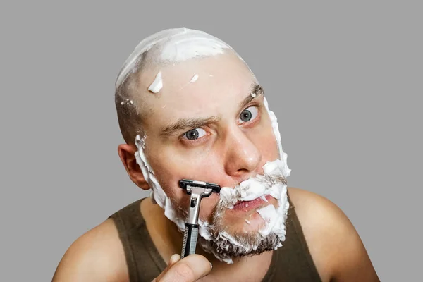 Un tipo con barba y una navaja en la mano, con espuma y gel en la cara se afeita por la mañana en casa —  Fotos de Stock