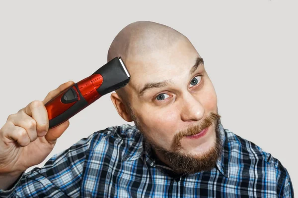 Bald guy with a beard and a clipper in his hands, shaving himself in the morning at home on a colored background