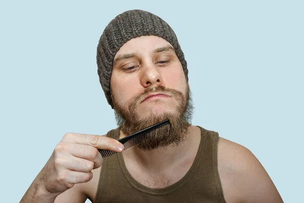 Close-up de um jovem estilo sua barba com um pente enquanto está sozinho em um estúdio contra um fundo azul — Fotografia de Stock