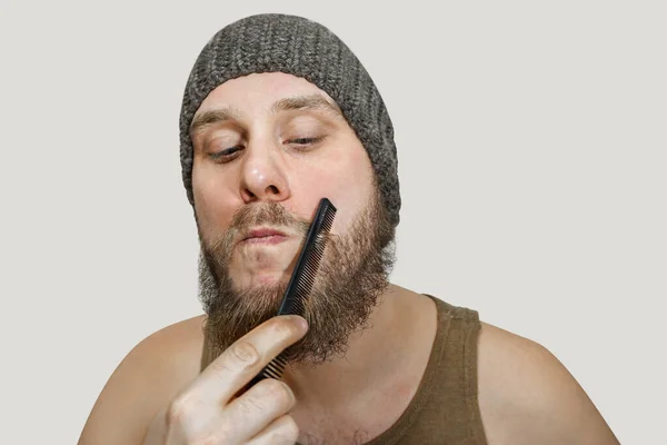 Retrato de um jovem barbudo de chapéu penteia a barba contra um fundo isolado — Fotografia de Stock