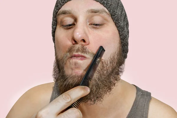 Портрет молодого бородатого парня в шляпе расчесывает бороду на изолированном фоне — стоковое фото
