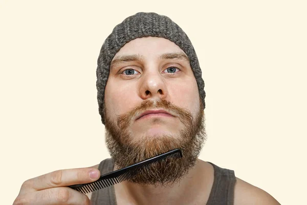 Retrato de um jovem barbudo de chapéu penteia a barba contra um fundo isolado — Fotografia de Stock