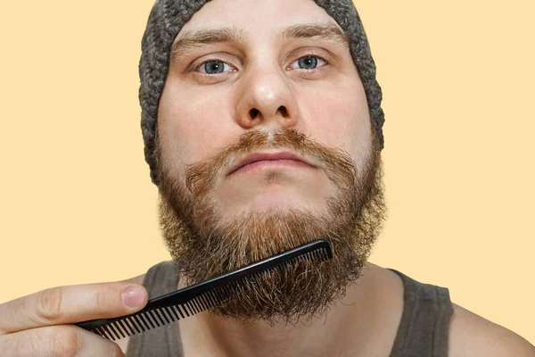 Portret van een jonge man met een baard in een hoed kamt zijn baard tegen een geïsoleerde achtergrond — Stockfoto