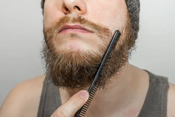 Portret van een jonge man met een baard in een hoed kamt zijn baard tegen een geïsoleerde achtergrond — Stockfoto