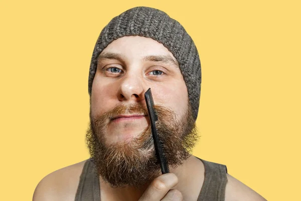 Portret młodego brodatego mężczyzny w kapeluszu grzebie brodę na odosobnionym tle — Zdjęcie stockowe