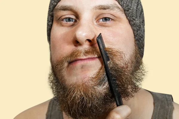 Retrato de um jovem barbudo de chapéu penteia a barba contra um fundo isolado — Fotografia de Stock