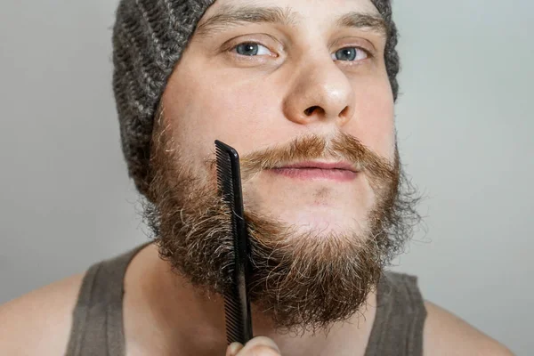 Portrét mladého vousatého chlapa v klobouku pročesává jeho vousy na izolovaném pozadí — Stock fotografie