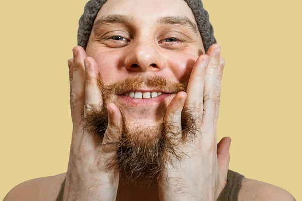 Retrato de um jovem barbudo de chapéu penteia as mãos de barba um fundo isolado — Fotografia de Stock