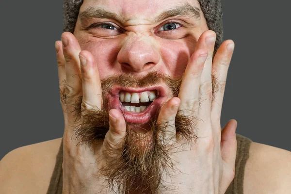 Портрет удивленного бородатого парня в шляпе с открытым ртом, расчесывающего бороду руками и пальцами в студии — стоковое фото