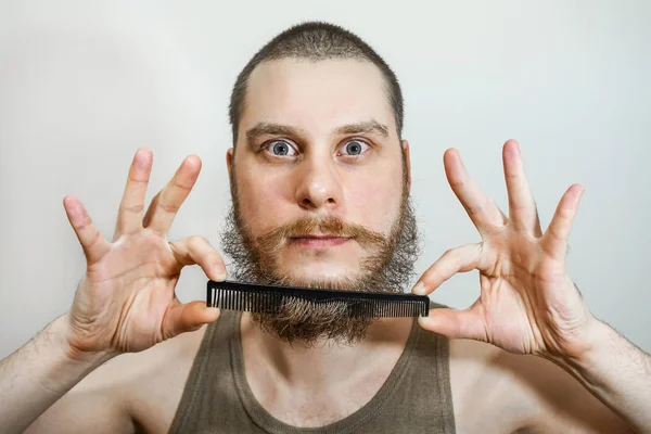 Primer plano de un joven que peina su barba con un peine mientras está solo en un estudio sobre un fondo gris — Foto de Stock