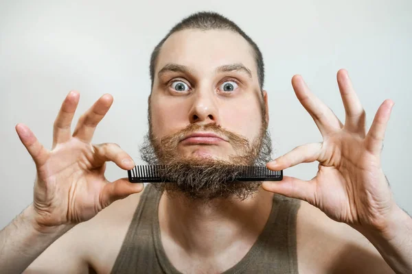 Close-up de um jovem estilo sua barba com um pente de pé sozinho em um estúdio contra um fundo cinza — Fotografia de Stock